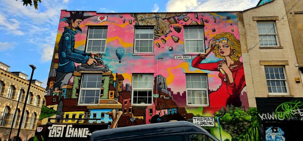 Mural by Silent Hobo on Stokes Croft showing a DJ at the decks, a Cheers Bristol balloon and a woman in red saying Come back