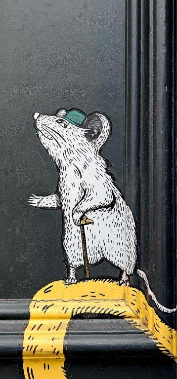 Small painted mouse in a cap leaning on a cane at the foot of a black door
