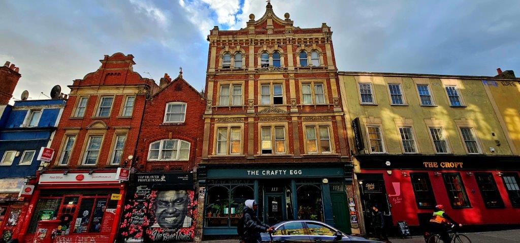 Victorian buildings on Stokes Croft housing the Post Office, The Crafty Egg and The Croft, with a We Save Lives mural on a shutter